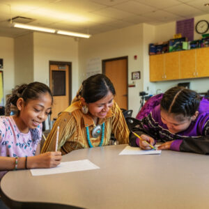 Two students work with an indigenous educator wearing traditional clothing and jewelry. 