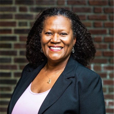 A woman with curly dark hair, wearing a black blazer over a light pink top, smiles in front of a brick wall background, embodying the spirit of the Antiracist Roadmap.