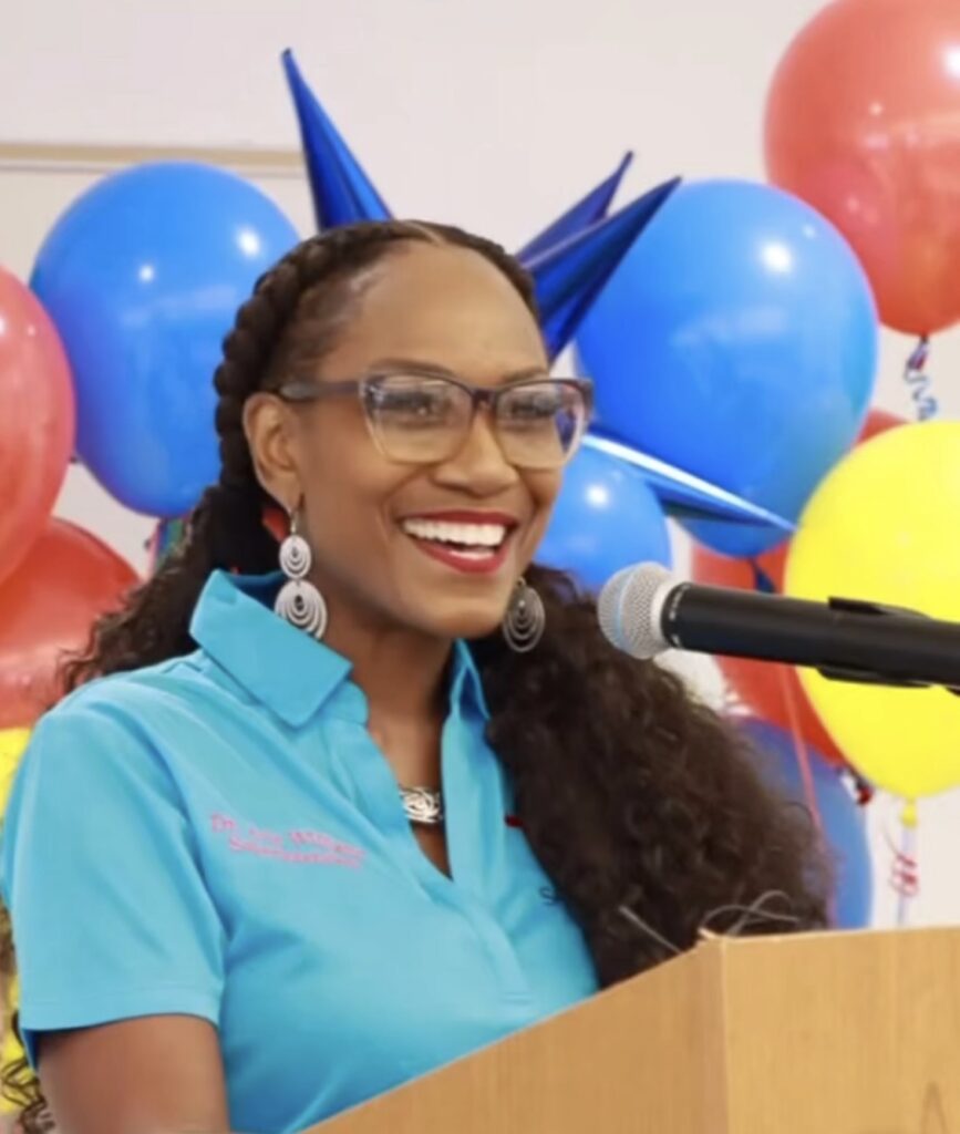 A photo of Dr. Avis Williams at a podium, in a bright blue shirt, with balloons behind her.