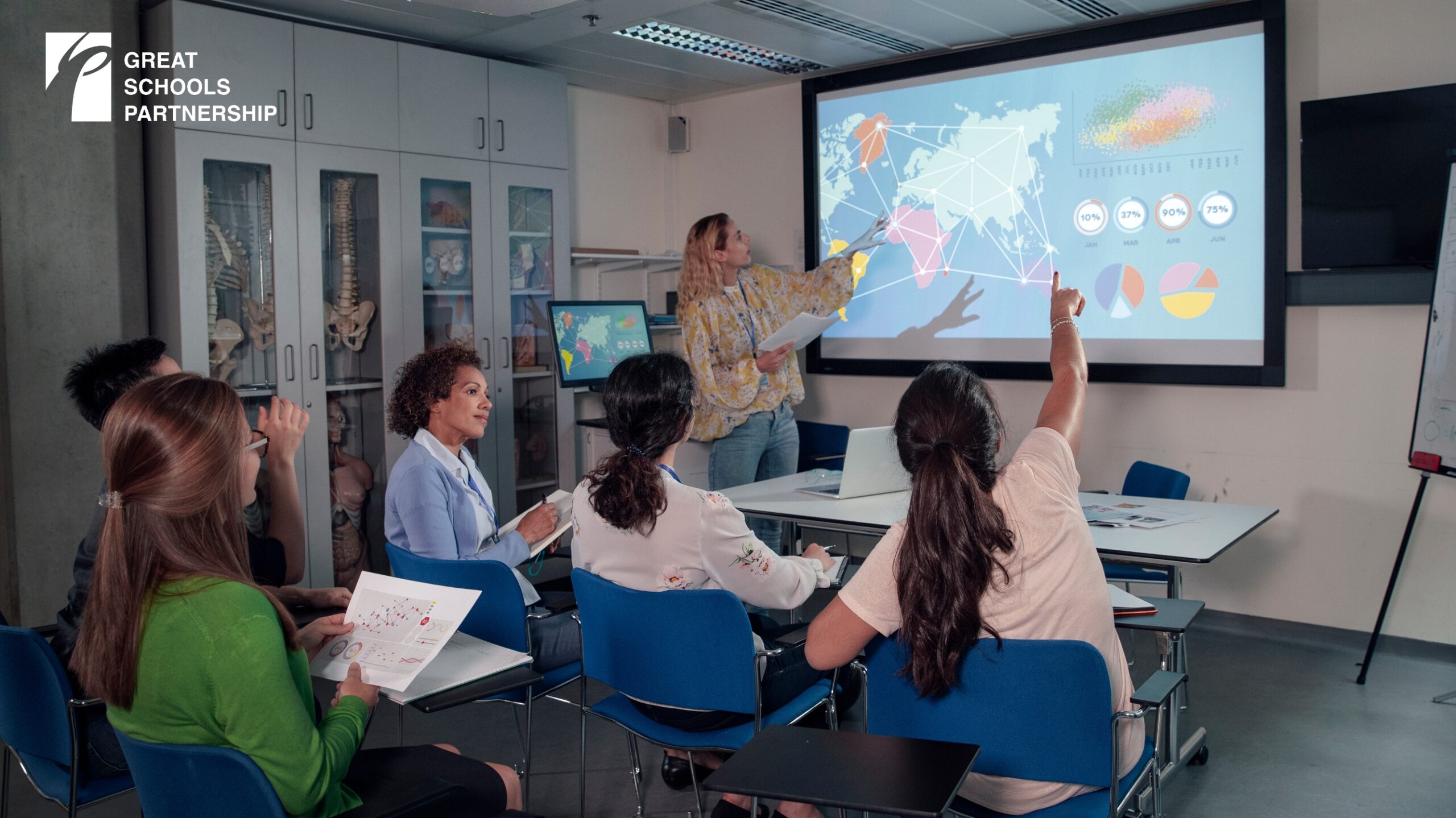 A teacher points to a board, while students raise their hands, and another educator looks on. 