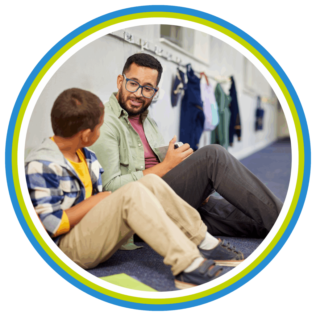An adult and a child sit on the floor, talking and smiling. The background shows hanging clothes, suggesting a school or casual setting. Both appear engaged in conversation, reflecting on their Antiracist Roadmap. The image is framed by colorful circular borders.