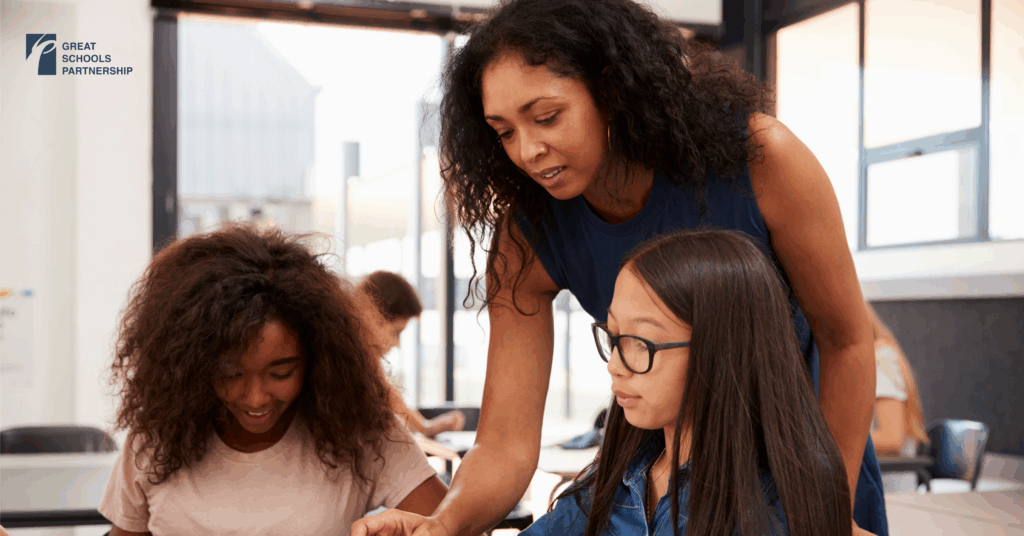 A teacher leans over a desk to help two students, one with curly hair and one with straight hair and glasses, in a bright classroom. The Great Schools Partnership logo is visible on the wall, highlighting that representation matters.