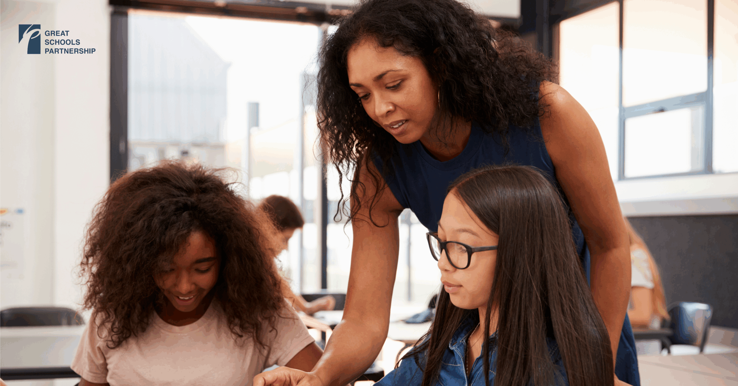 A teacher leans over a desk to help two students, one with curly hair and one with straight hair and glasses, in a bright classroom. The Great Schools Partnership logo is visible on the wall, highlighting that representation matters.