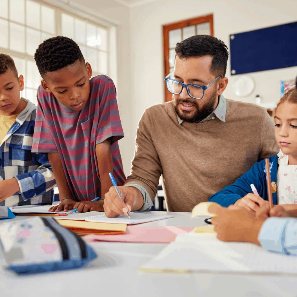 Dr. Lamirra Hood-Stewart, a champion of representation in education, helps a group of young students with their schoolwork at a classroom table. The children are focused on writing, with books and supplies spread out before them.