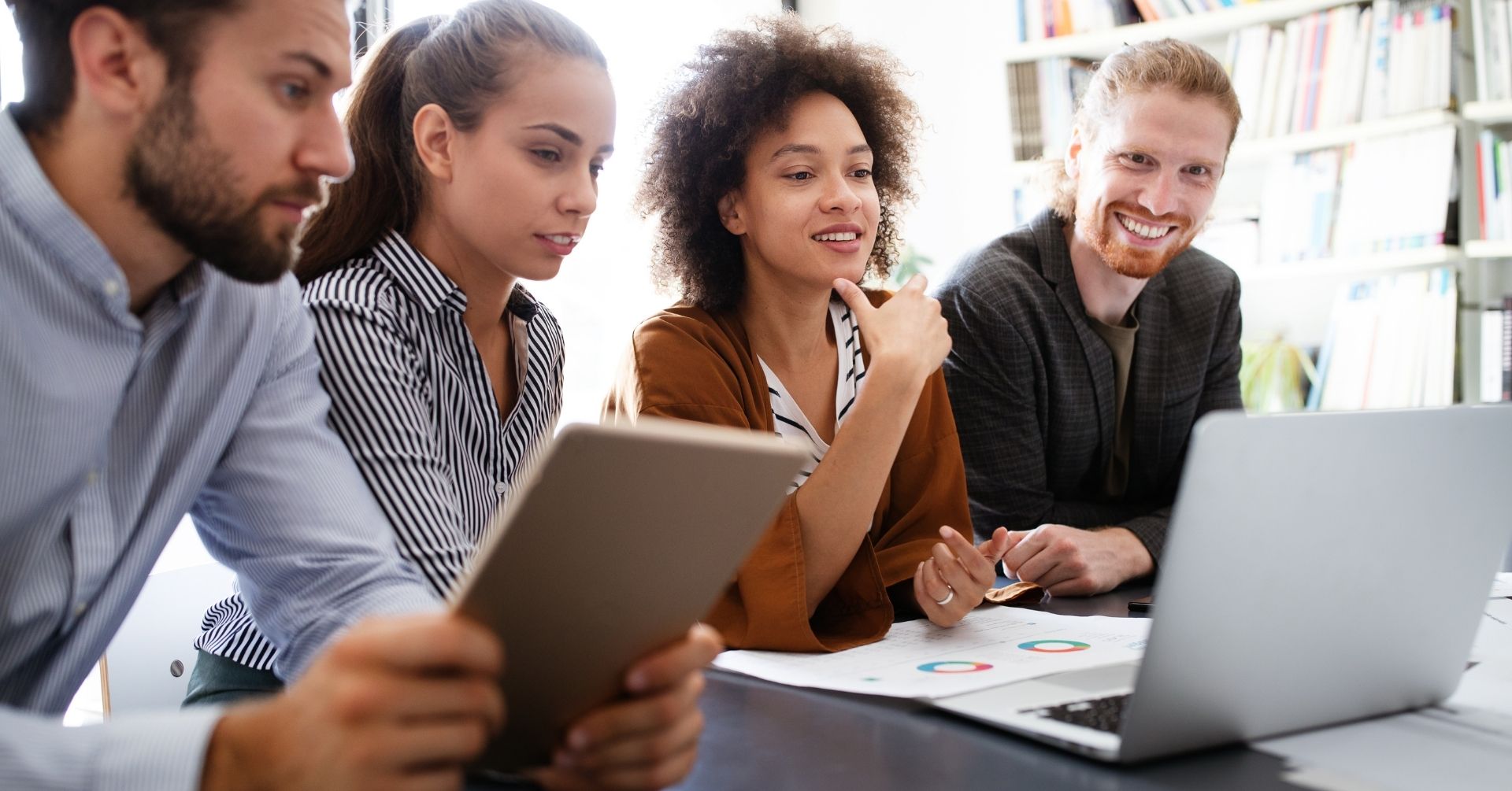 Four people sit at a desk collaborating, with one using a tablet and others looking at a laptop. Charts and papers fill the table as this New England Community Consortium team appears engaged and focused in a bright office setting.