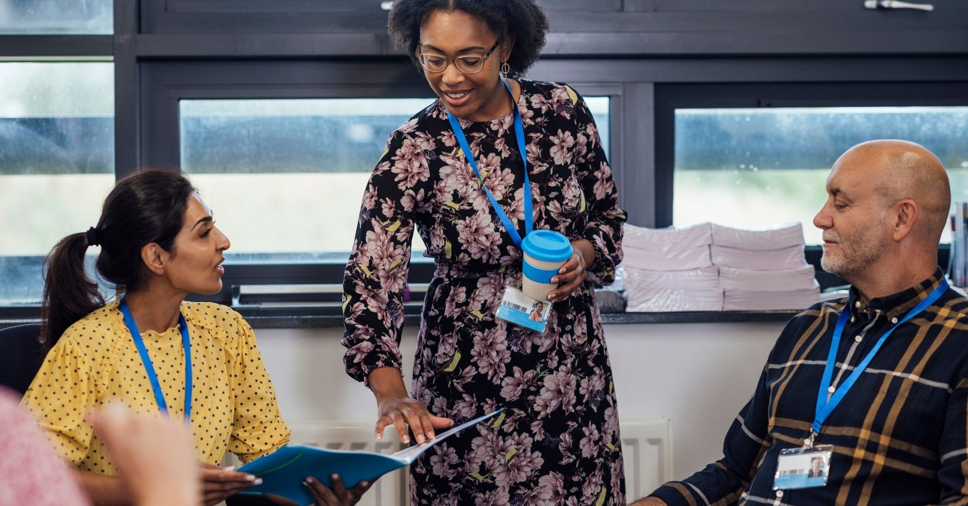 Three adults in a New England District Community Consortium meeting room, wearing ID badges, engage as one woman in a floral dress stands with a cup, pointing at papers while the other two listen attentively.