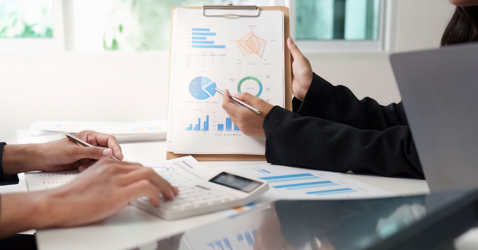 Two people from the District Community discuss business charts and graphs on a clipboard at a desk. One points with a pen, while the other uses a calculator. Papers and a laptop representing their consortium are also visible on the table.