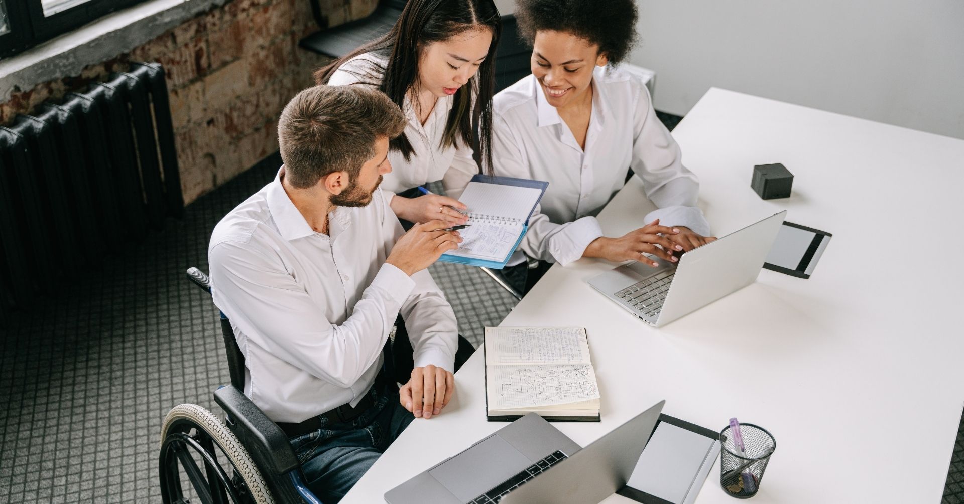 Three colleagues, one using a wheelchair, collaborate at a white table with laptops and notebooks, engaged in discussion and planning for a District Community Consortium in a modern office setting.