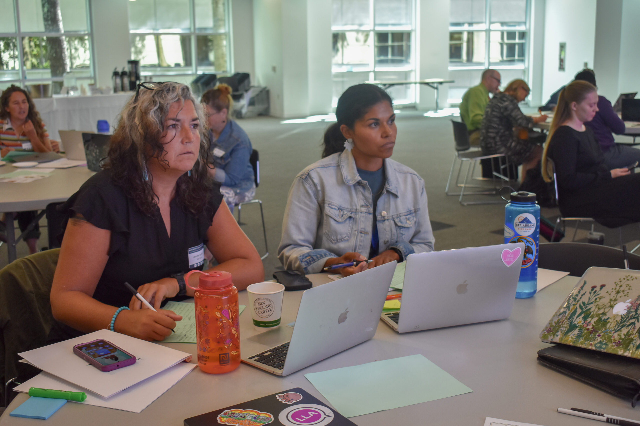 Two women sit at a table with laptops and notebooks, attentively listening in a bright, modern classroom with large windows—a dynamic setting fostering Maine Education as other groups work and converse in the background.