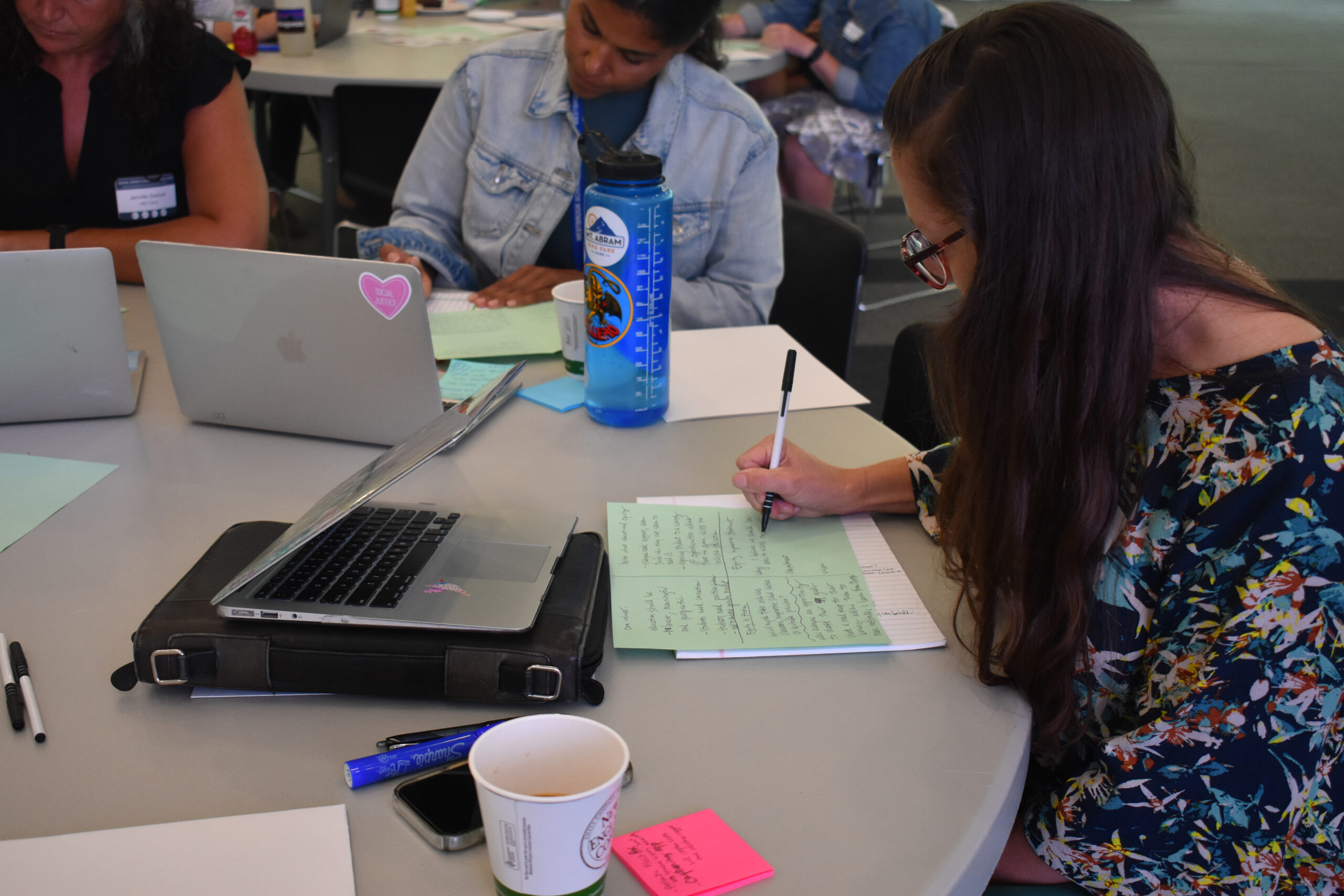 Three people sit at a round table with laptops, notebooks, and green paper, writing notes. A large water bottle and coffee cup sit nearby. The setting appears to be a Maine Impact Education Fellowship group work session.