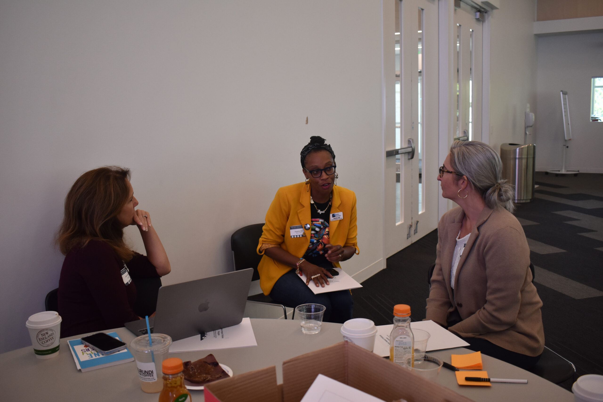 Three women sit around a table engaged in conversation, likely discussing their Maine Impact Education Fellowship. Papers, drinks, snacks, and a laptop are scattered across the table in their meeting room.