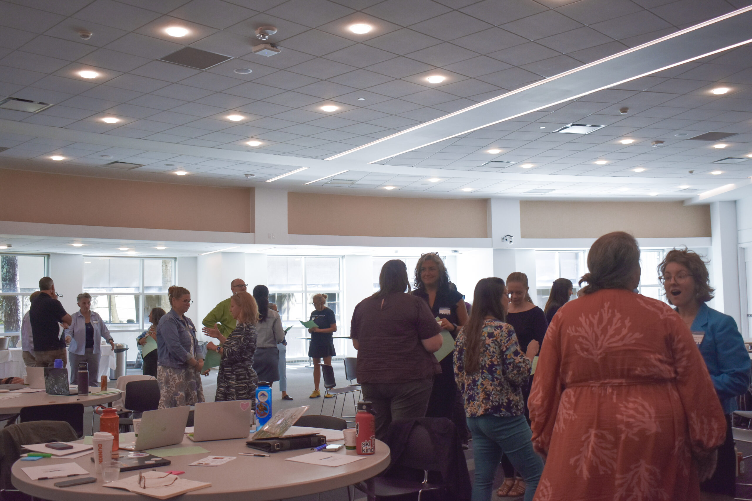 Attendees of the Maine Impact Education Fellowship gather in a brightly lit conference room with round tables and large windows, holding notebooks and drinks as they engage in lively conversation.