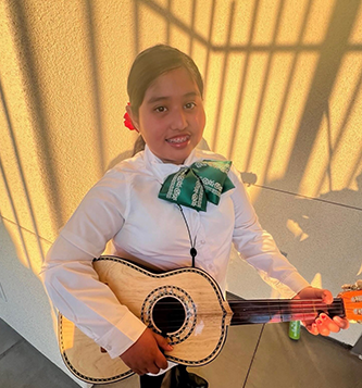 A young child wearing a white long-sleeved shirt and green bow tie smiles while holding a guitar in a sunlit room.