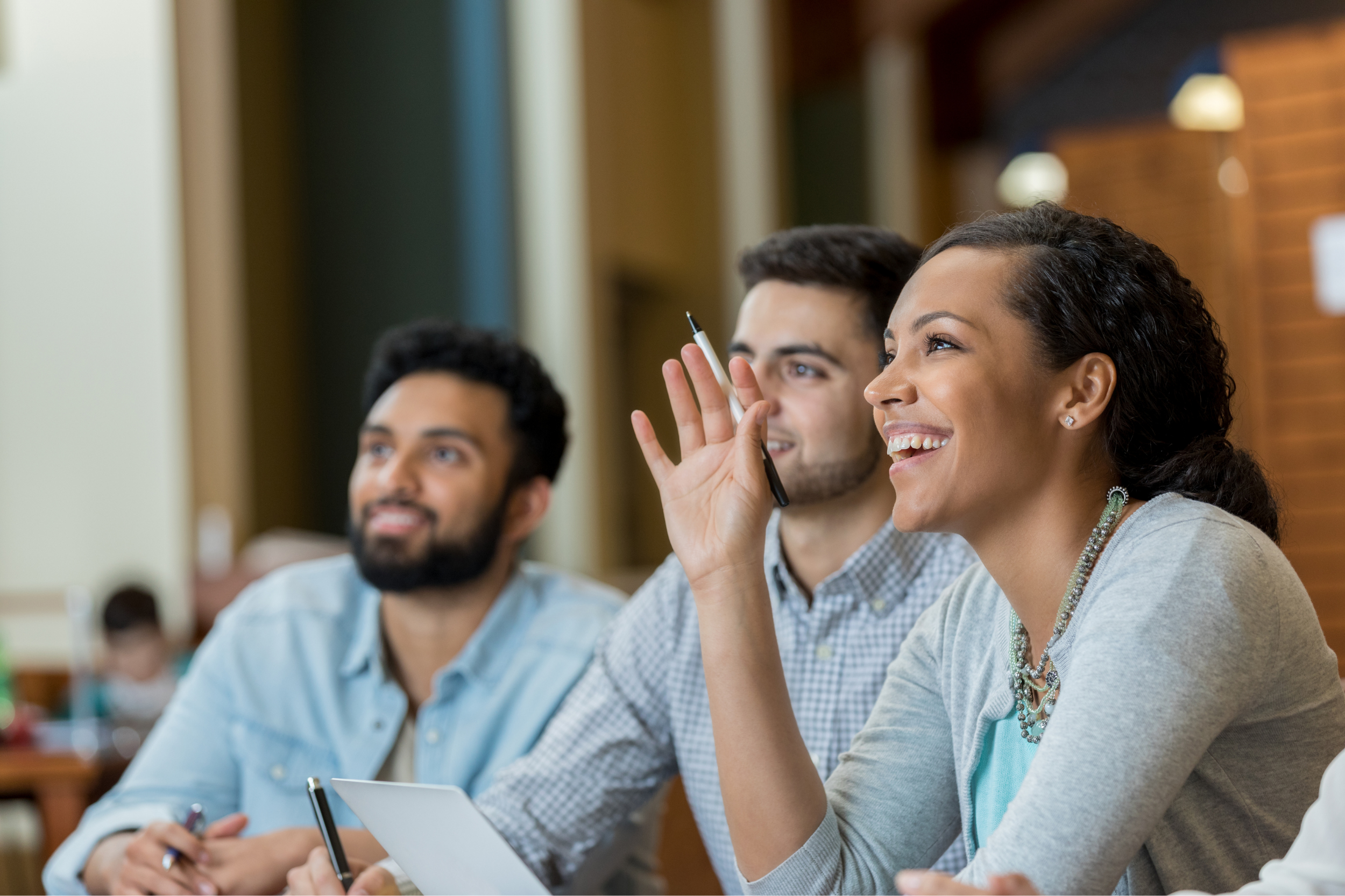 Three adults sit together in a classroom. A smiling woman raises her hand, while two men beside her look forward attentively. Holding pens and notebooks, they appear engaged in Discovery Sessions, actively participating in a discussion or lesson.