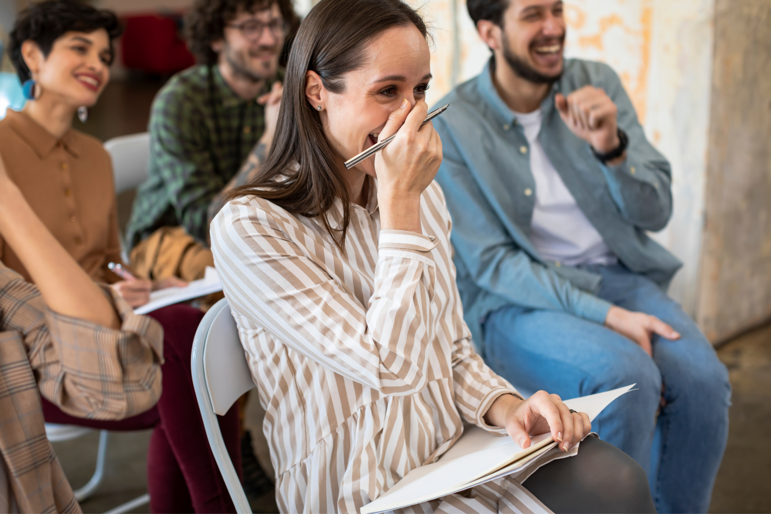 A group of people sitting indoors on chairs, laughing and smiling. A woman in the foreground covers her mouth with her hand while holding a notebook and pen, appearing amused during Discovery Sessions or a workshop.