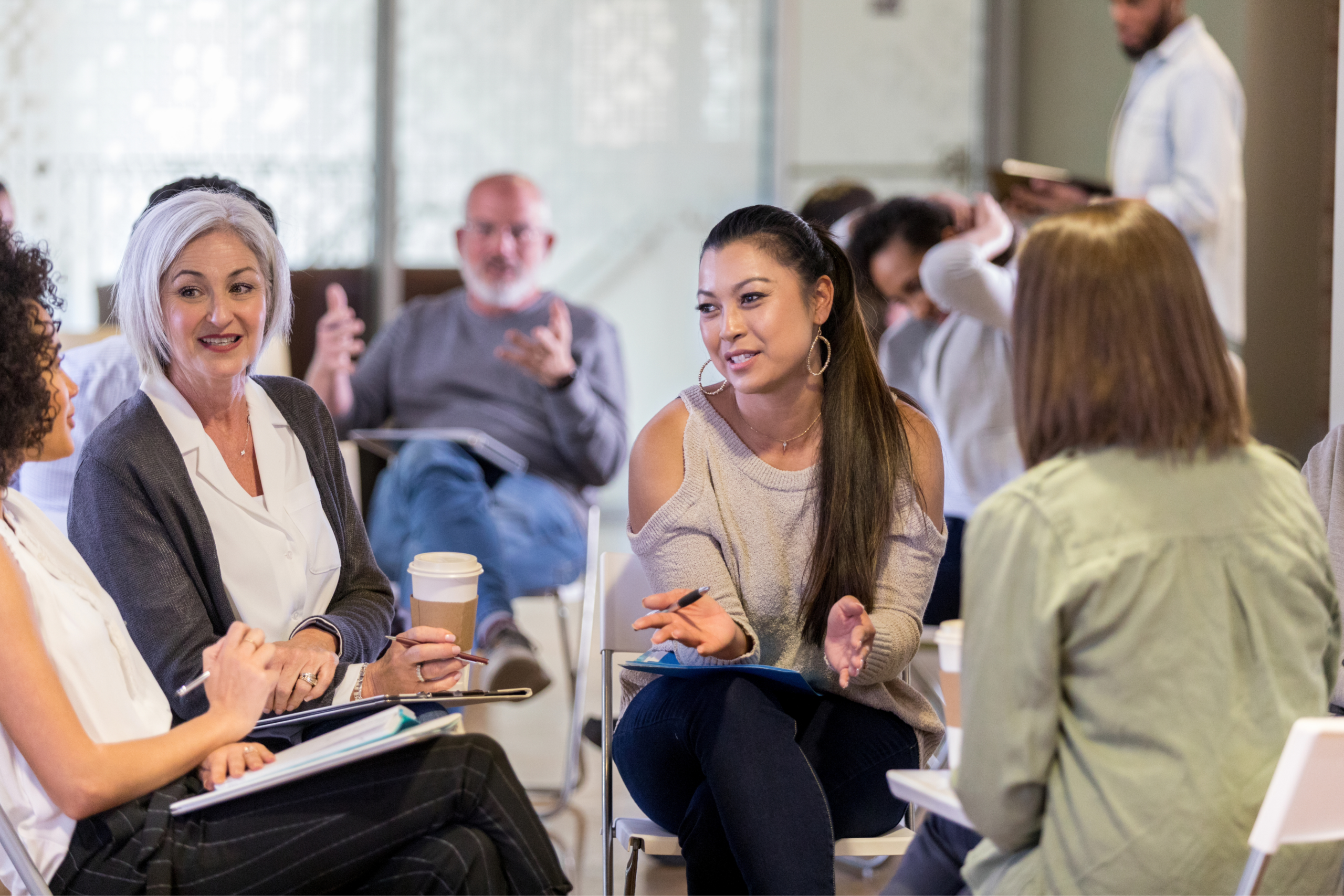 A diverse group of people sit in a circle, engaged in Discovery Sessions. Some hold notebooks and coffee cups, suggesting a casual meeting or discussion in a bright, modern room.