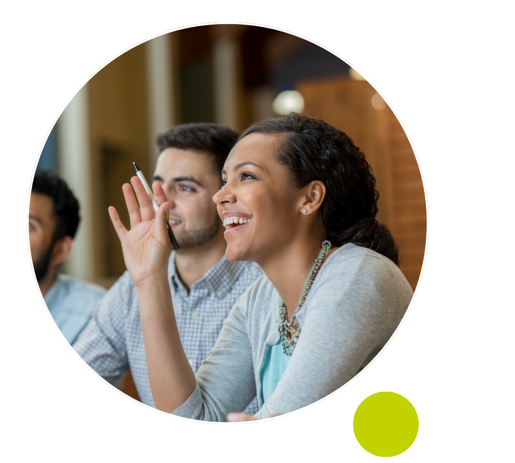 A woman smiling and raising her hand with a pen, seated beside a man during Discovery Sessions in a classroom or meeting setting. The image features a circular frame with dotted design elements.