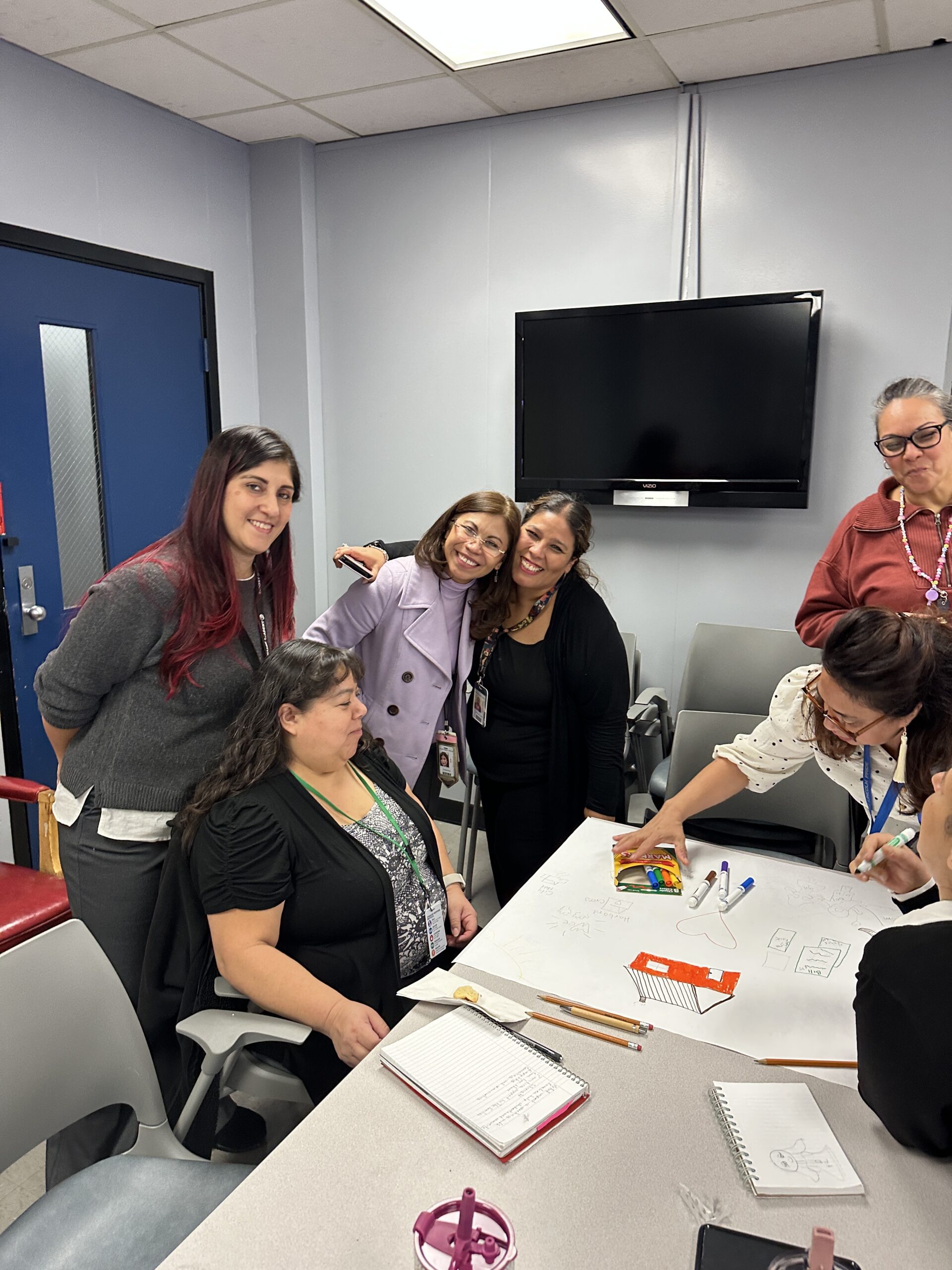 A group of six women gathers around a table in an office, collaborating on Family Engagement in Schools. Some are standing, some seated, with a few smiling at the camera while others draw with colored markers. A TV is mounted on the wall behind them.