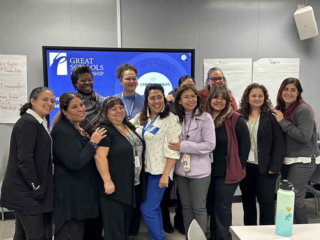 A group of thirteen smiling adults stand together in a classroom, posing for a photo in front of a screen displaying “Great Schools Partnership,” highlighting their commitment to Family Engagement in Schools. Flip charts and a water bottle are visible in the background.