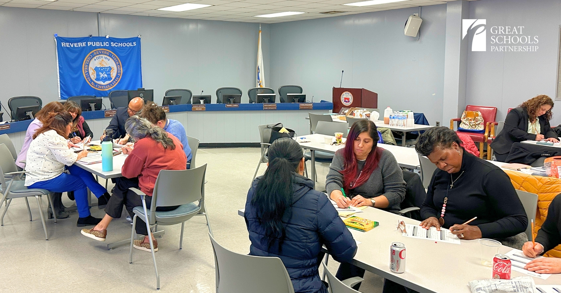 Adults sit in groups at tables, writing on papers in a conference room with a Revere Public Schools banner and Great Schools Partnership logo visible, collaborating to strengthen Family Engagement in Schools.
