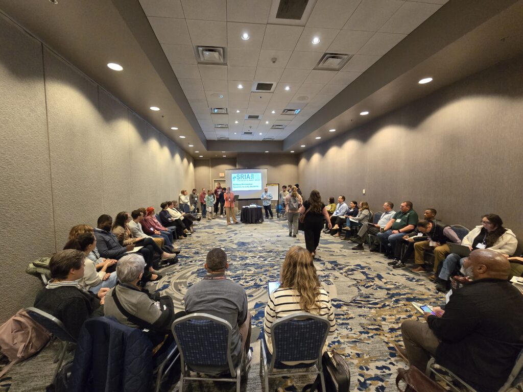 A large group of people sits in a circle in a conference room during the conference, while a few individuals stand at the front near a screen displaying the PSAAL logo. The atmosphere appears to be collaborative and attentive.