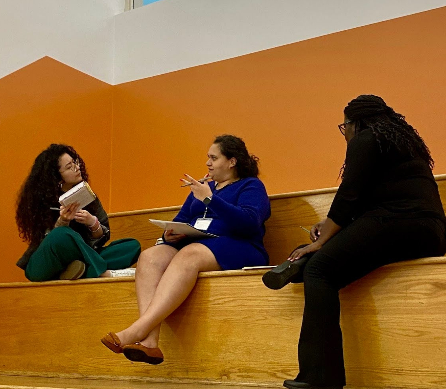 Three women sit on wooden steps against an orange wall, engaged in collective learning. One writes in a notebook while the others listen and gesture, all focused and involved in discussion within this safe learning space.