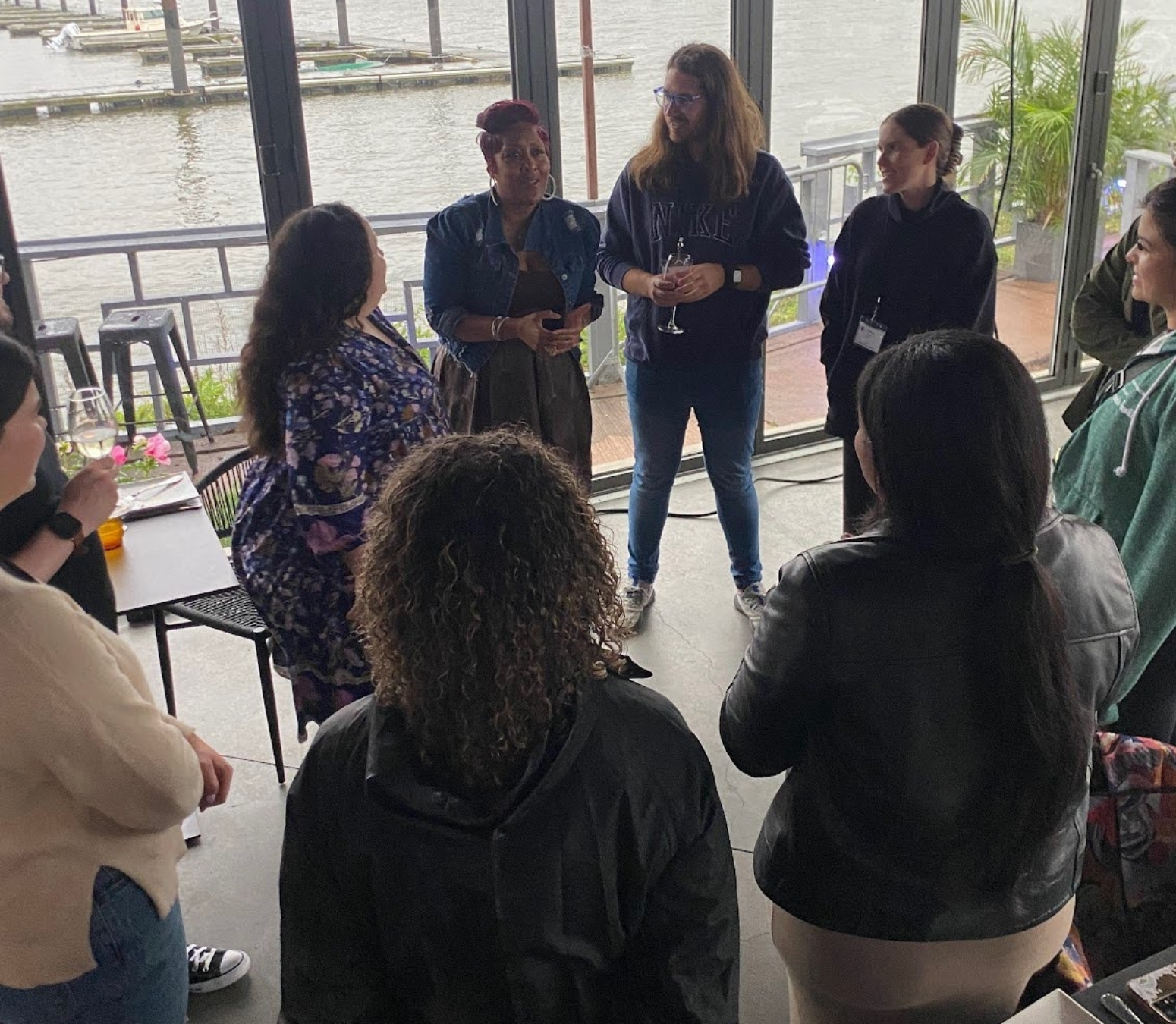 A group of people, including school leaders, stand in a circle indoors near large windows overlooking water. They appear engaged in conversation within collective learning spaces, some holding drinks, and a marina visible outside.