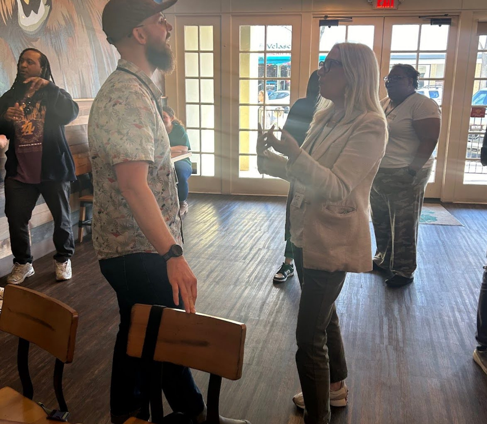 A woman and a man stand facing each other and talking in a sunlit room with wooden floors, as school leaders foster collective learning. Three others are in the background near large windows and doors.
