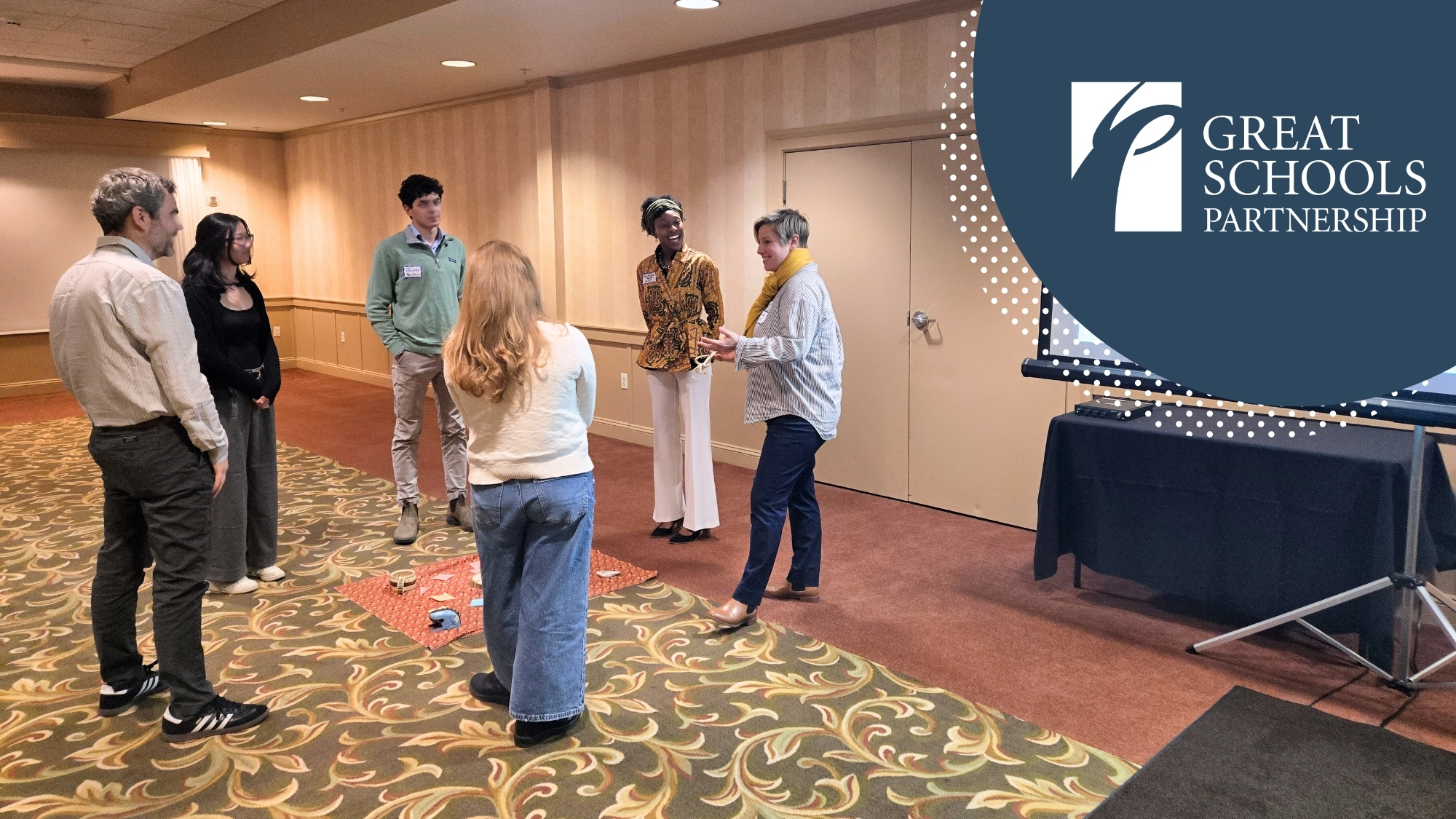 Six conference participants stand in a small circle in a hotel ballroom, engaged in a facilitated activity. Materials are spread on a mat on the floor in the center of the group. The Great Schools Partnership logo appears in the upper right corner of the image against a dark teal background with a dotted pattern.