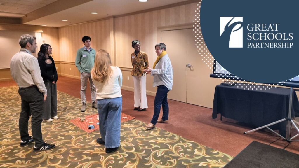 A group of six adults stands in a circle in a conference room, attentively listening to a woman speaking during the conference. A screen displaying the Great Schools Partnership logo is visible to the right.