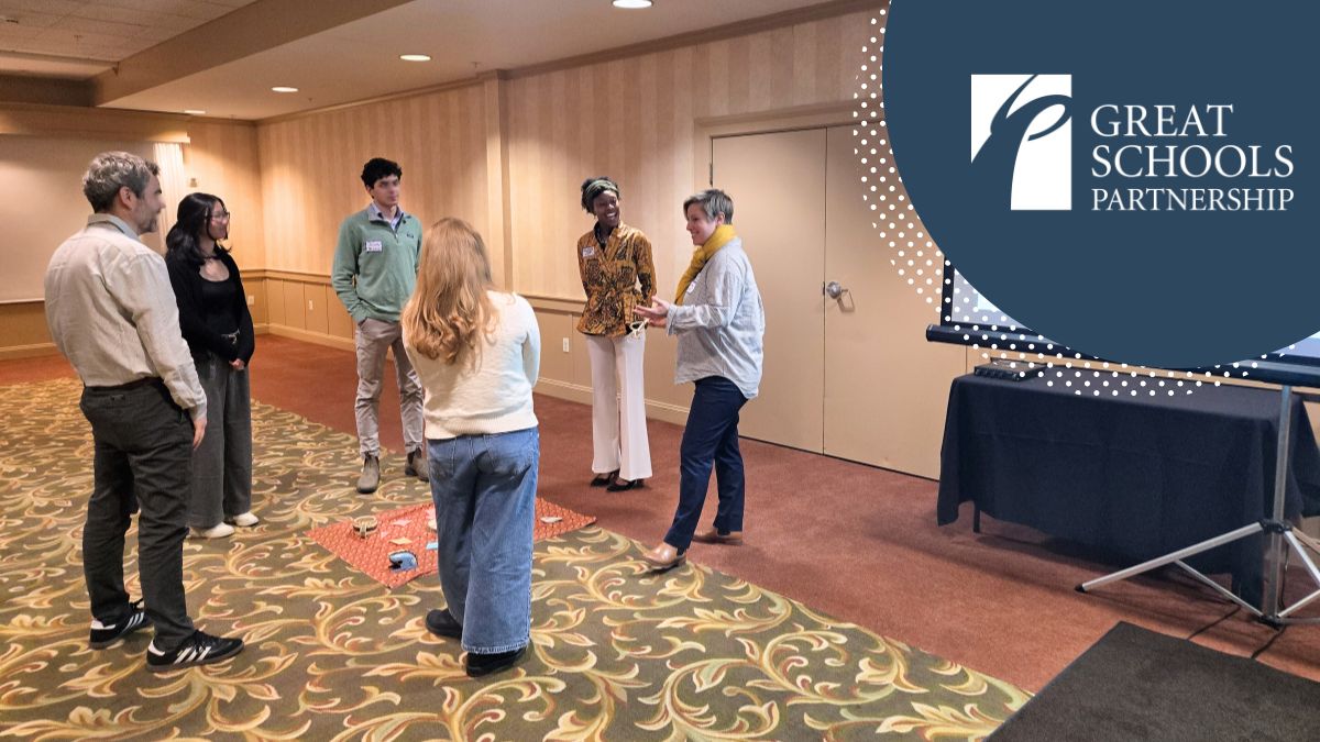 A group of six adults stands in a circle in a conference room, attentively listening to a woman speaking during the conference. A screen displaying the Great Schools Partnership logo is visible to the right.