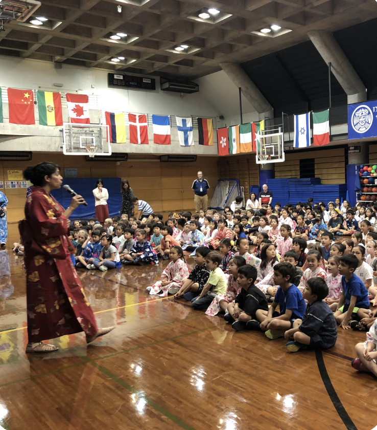 A woman in a red kimono speaks to a large group of children seated on a gym floor in one of the school’s collective learning spaces. Various national flags hang above, and several adults, including school leaders, are present in the background.