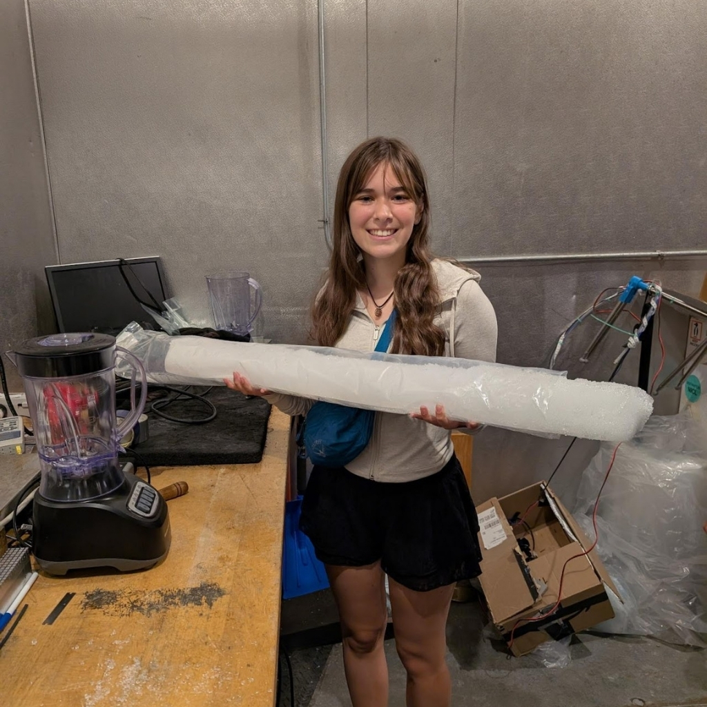 A young woman smiles while holding a long cylinder of ice beside a blender on a workbench in a classroom with metal walls and various equipment, showcasing hands-on lessons in an industrial setting.
