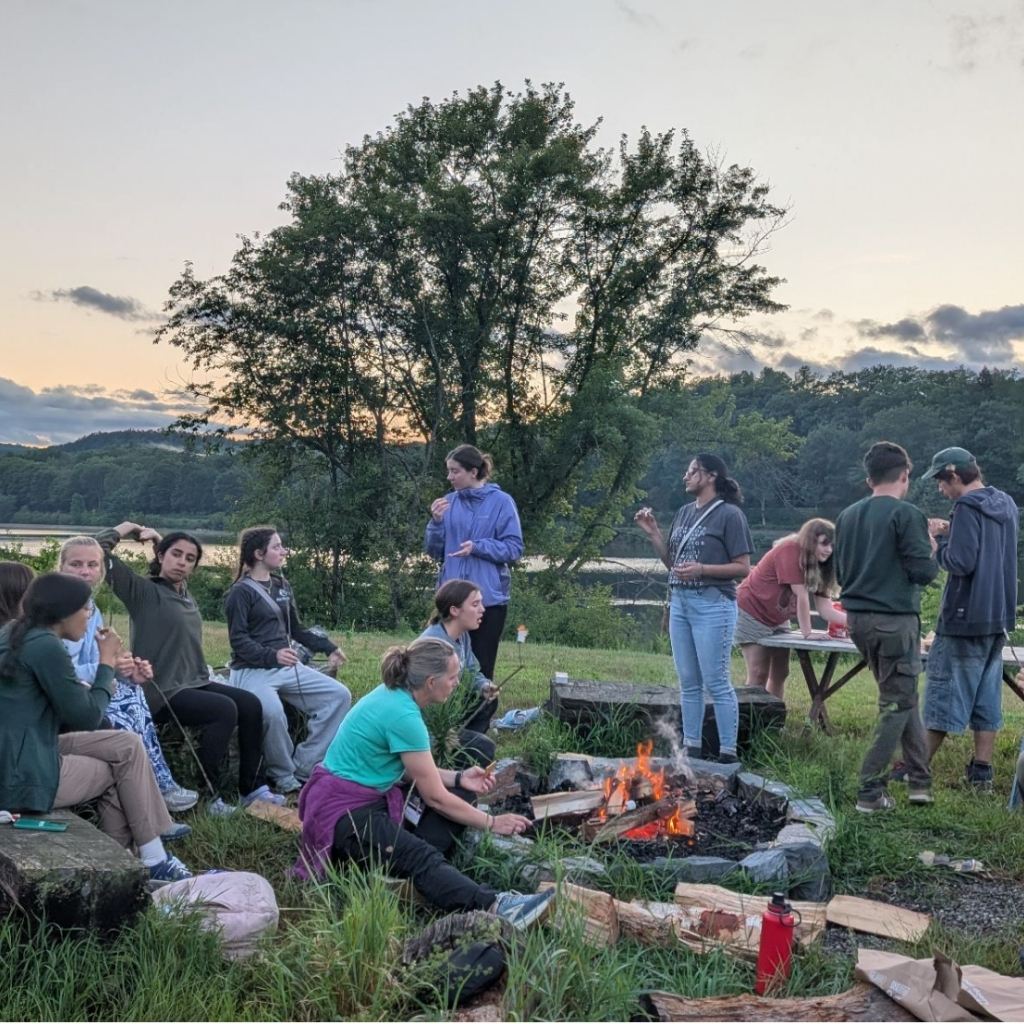 A group of people sit and stand around a campfire outdoors at sunset, with a lake and trees in the background. Some are talking, roasting marshmallows, or preparing food—turning the moment into an outdoor learning classroom of shared lessons and laughter.
