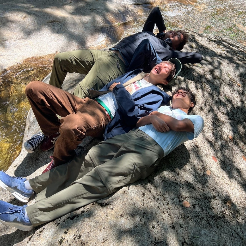 Three people are lying on a large rock outdoors, enjoying the sunlight as if their classroom has shifted outside. They appear relaxed, smiling with their eyes closed, surrounded by dappled shadows from nearby trees.