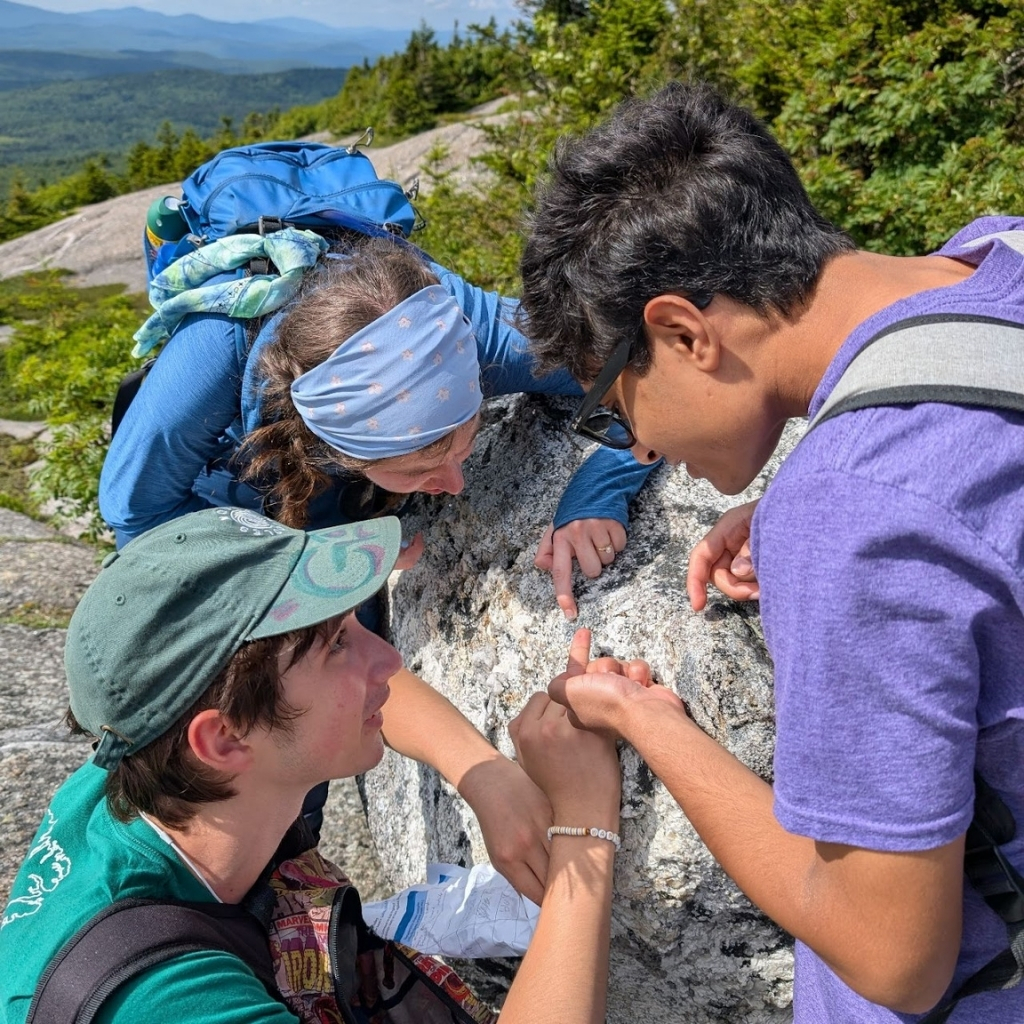 Three people engage in outdoor learning on a large rock while hiking. One points at the rock, another looks through glasses, and the third kneels beside them. Green trees and distant mountains frame this classroom without ceiling.