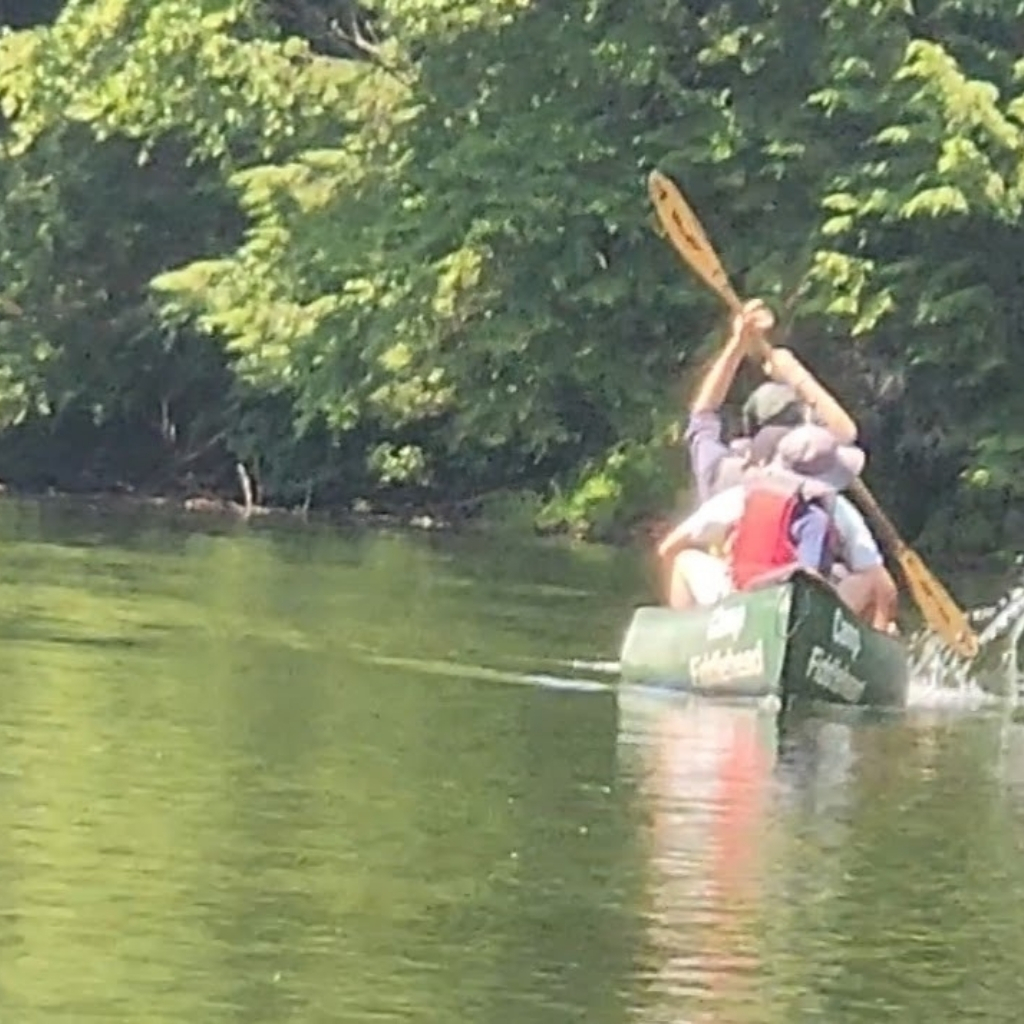 Two people paddle a green canoe down a calm river, surrounded by lush green trees on a sunny day, enjoying outdoor learning far beyond the classroom.
