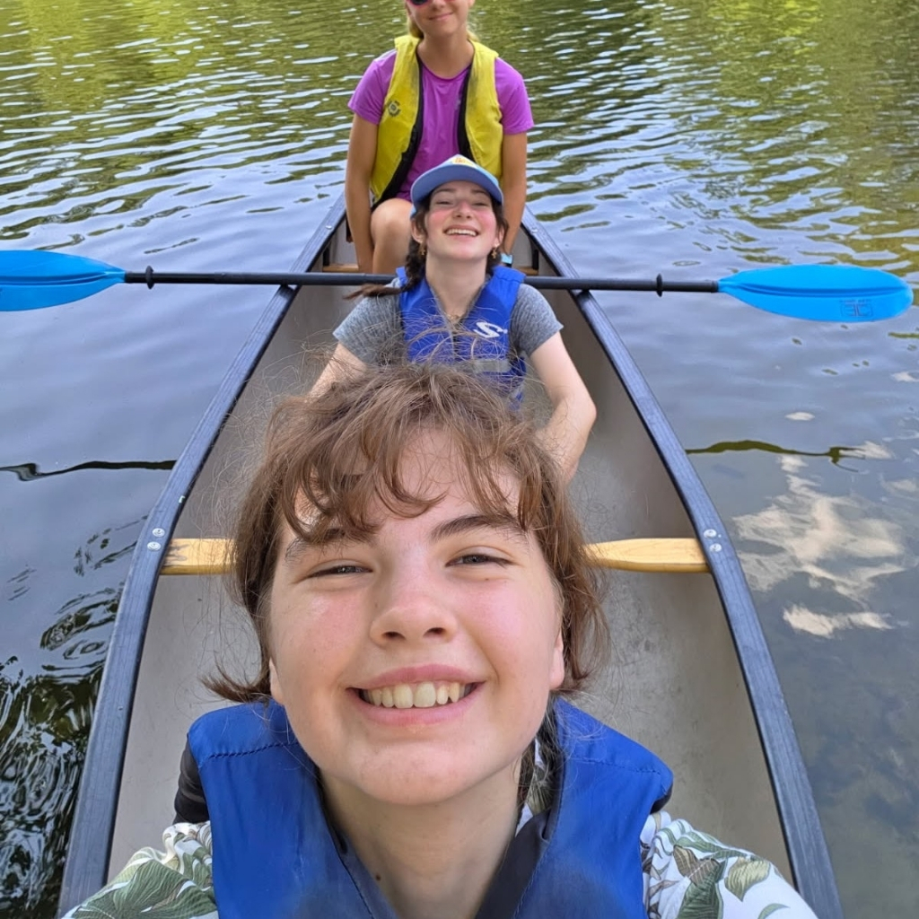 Three smiling people in life jackets sit in a canoe on calm water, with paddles resting on the sides. The person in front takes a selfie, while the others enjoy outdoor learning and lessons in nature on a sunny day.