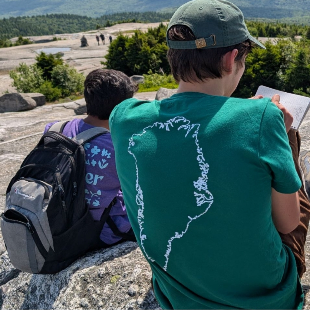Two people sit on rocky ground facing away from the camera, turning nature into their classroom. One in a green shirt with an outline of Greenland writes lessons in a notebook; the other, in purple with a black backpack, enjoys outdoor learning among trees and mountains.