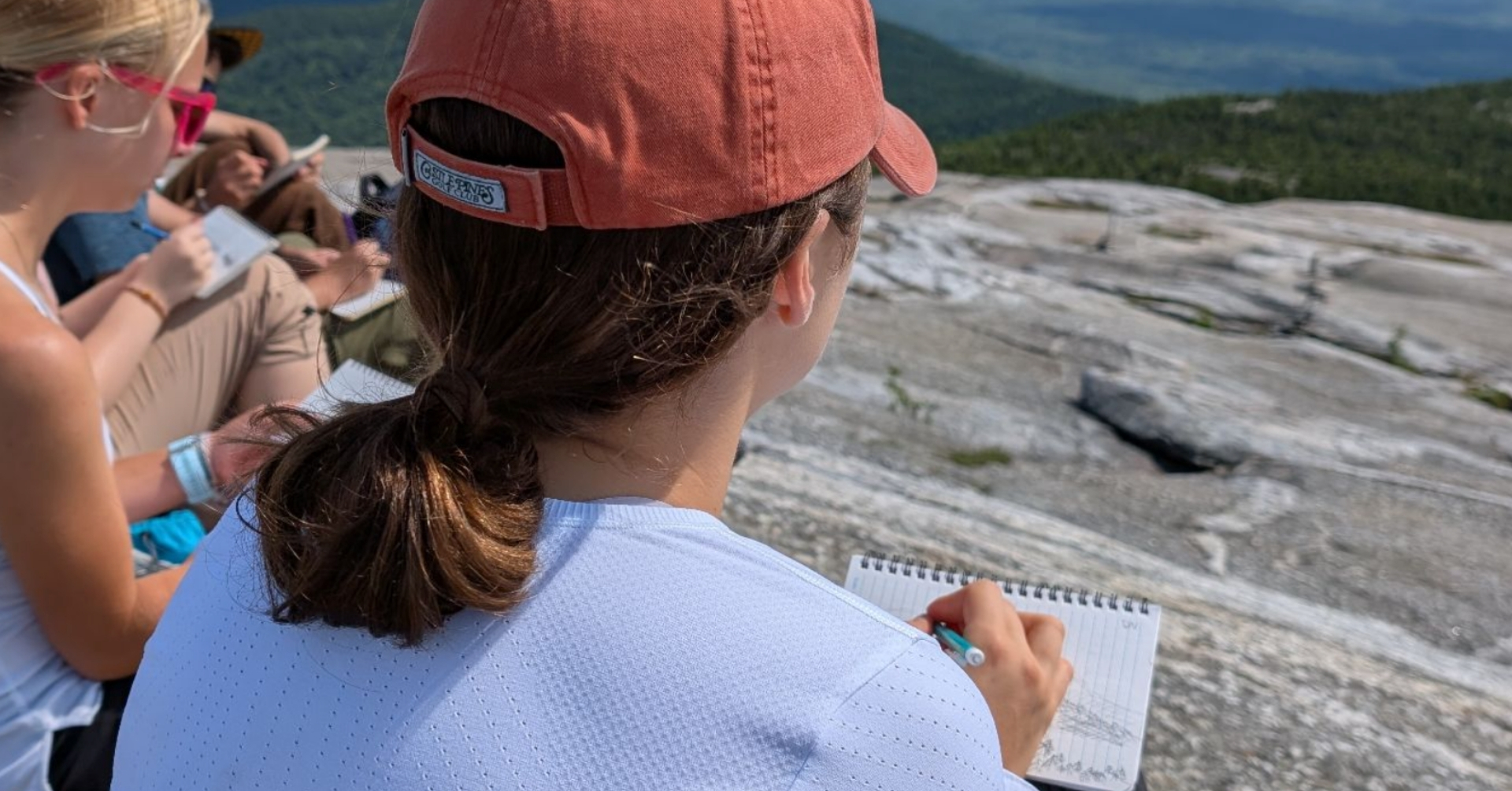 A person wearing a red cap and white shirt sketches in a notebook while sitting on a rocky mountain slope, embracing outdoor learning as another person looks on and a scenic, green landscape stretches out in the background.