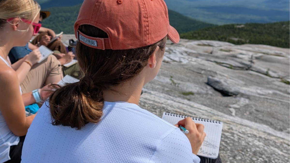 A person in a red cap sketches in a notebook while sitting on a rocky mountaintop with others, embracing outdoor learning. The background reveals forested hills and a vast landscape under a clear sky—a true classroom without ceiling.