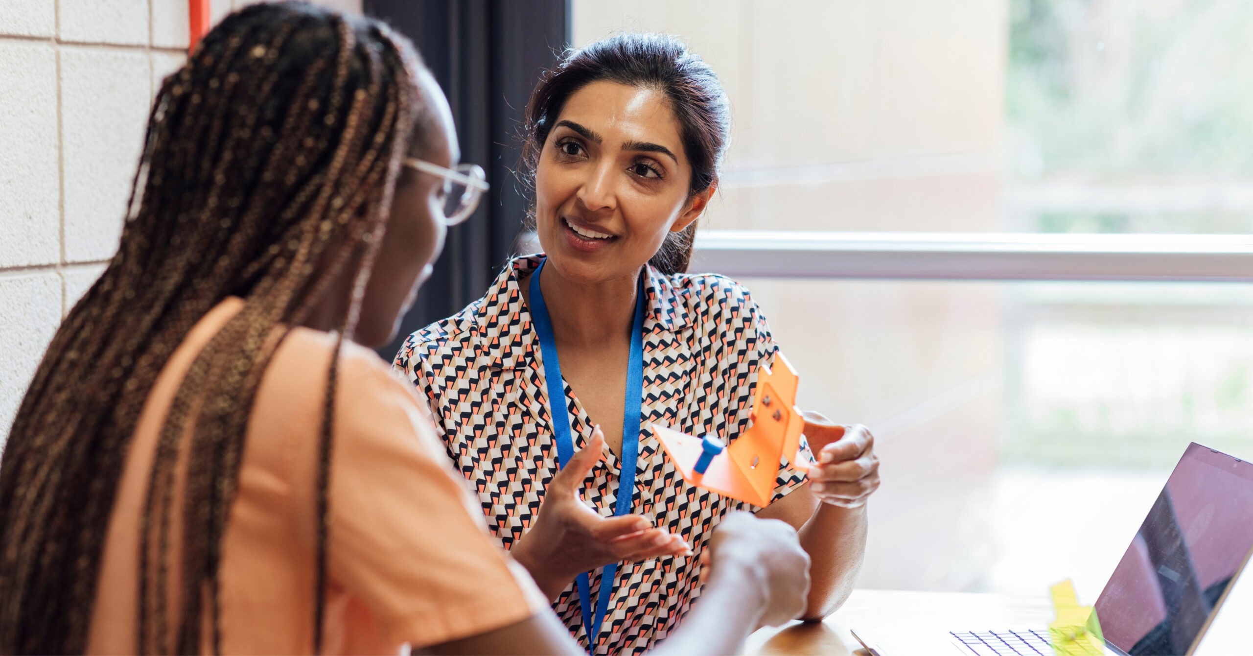 Two women sit at a table in discussion. One woman holds an orange geometric paper shape and gestures while speaking about Proficiency-Based Learning. The other listens attentively. A laptop and notebook are on the table as sunlight streams through a window.