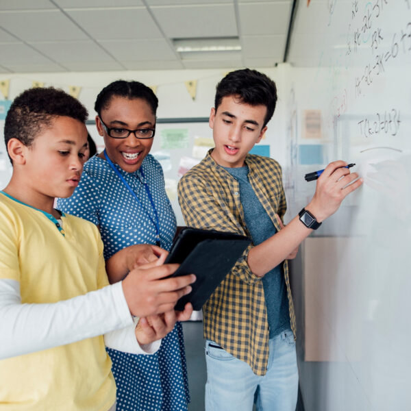 Three students stand by a whiteboard; one writes equations, another holds a tablet, and a third smiles while looking on. They appear to be collaborating on a math problem using Proficiency-Based Learning in a classroom.