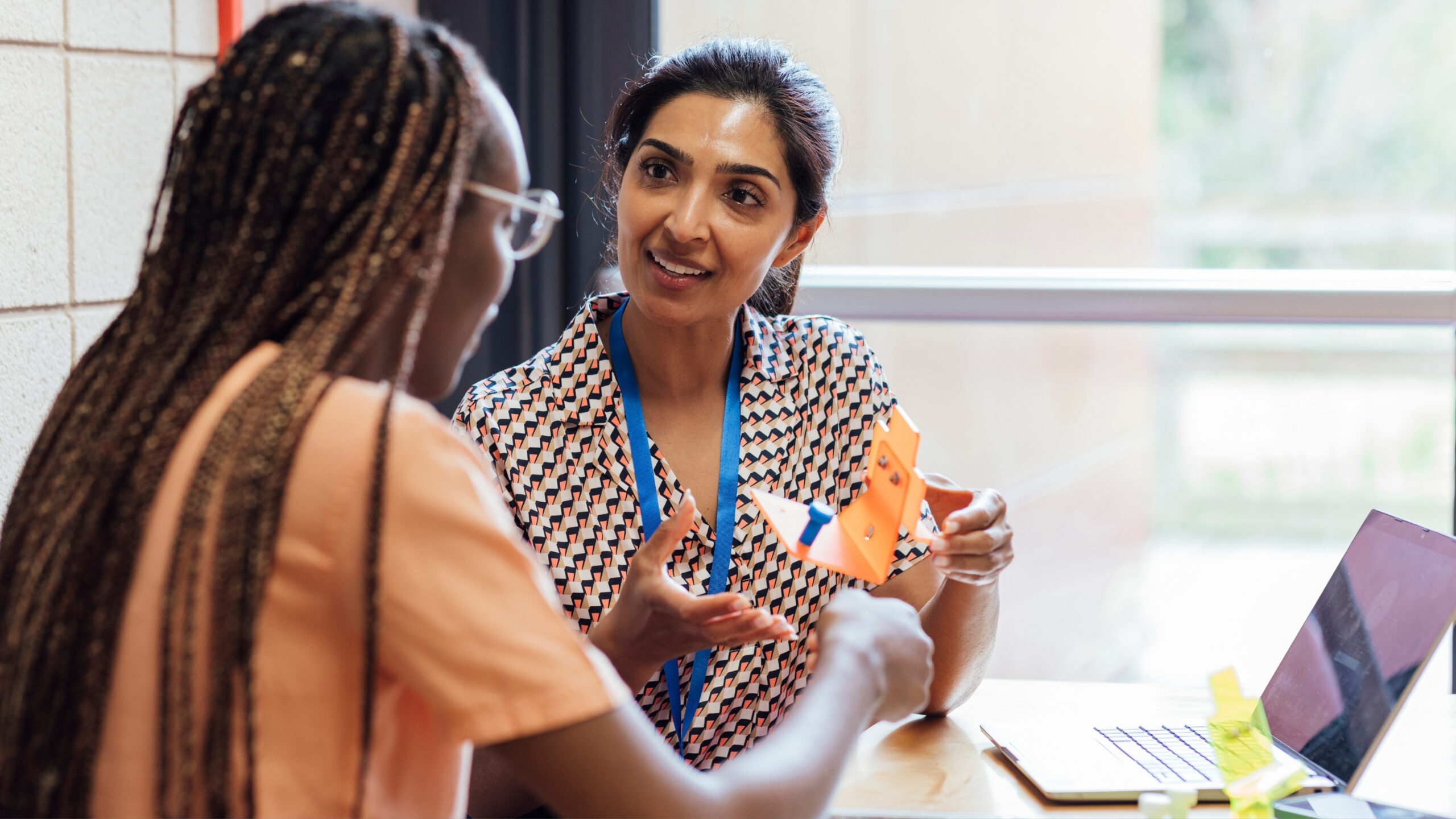 Two women sit by a window, engaged in a collaborative conversation about Proficiency-Based Learning. One holds an orange origami figure as they discuss ideas, with a laptop covered in sticky notes open in front of them.