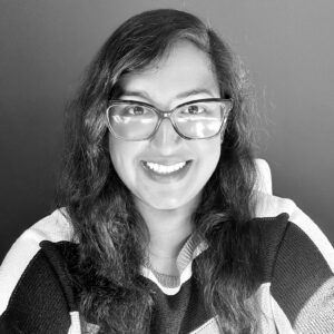 Black and white photo of a person with long wavy hair and glasses, smiling at the camera. Wearing a striped sweater, they sit in front of a plain dark background—capturing the thoughtful spirit behind Proficiency Based Learning.